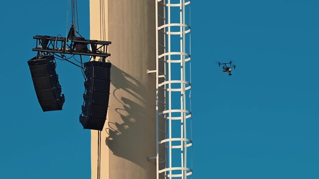 Professional drone flying near vertical industrial tower with hanging audio speakers and ladder