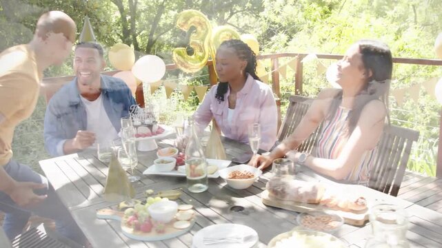Vertical video: Leaning friend in orange shirt placing hat on denim celebrant on deck, toasting 30