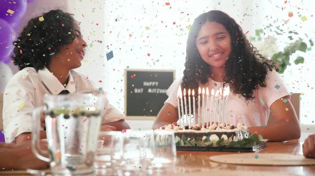 Teenage girl making wish and blowing candles on big cake at family table, causing confetti overlay