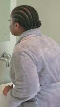 Vertical video: Turning lady with braids in grey robe placing pink eye patches at sink for eye care