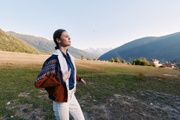 Naklejka premium Woman in nature standing in an open field beneath distant mountains, eyes closed breathing deeply, casual portrait wearing a chunky sweater and scarf, peaceful relaxation outdoors.