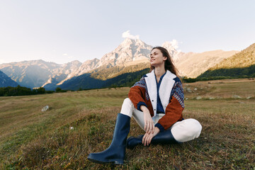 Naklejka premium Woman sitting on meadow in front of mountain landscape, outdoor portrait of young woman wearing jacket and boots, relaxed pose in nature with scenic horizon and clear sky.