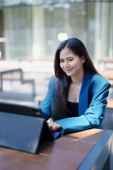 Obraz premium Young Asian businesswoman smiling while typing on a digital tablet at a sunny outdoor café, illustrating remote work, flexible lifestyle, professional connectivity and productivity