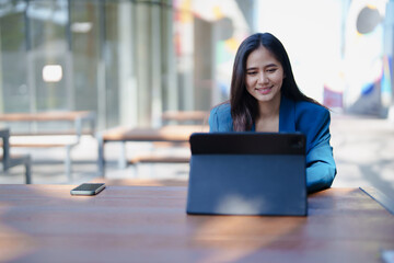 Young Asian businesswoman smiling while focusing on her digital tablet, working remotely from an...