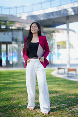 Obraz premium Asian business woman standing outdoors holding a laptop, wearing a pink blazer and white pants, looking away with a confident smile against an urban background