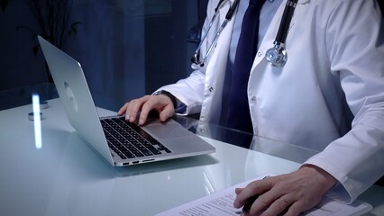Medical doctor in lab coat with stethoscope typing on laptop in a dim office, managing electronic...