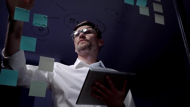 Man in eyeglasses organizing thoughts and ideas for business optimization on a clear glass board. Creating a strategic roadmap for innovation and project planning