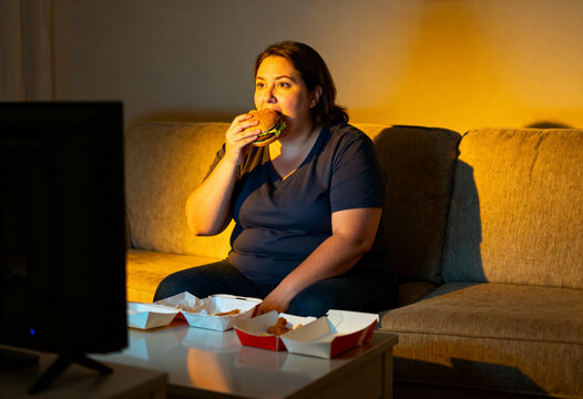 Woman eating fast food burger while watching television on sofa at night. Overweight person having impulsive midnight snack. Unhealthy lifestyle, dietary habit, binge eating concept.