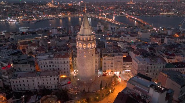 Morning view of Galata Tower illuminated against historic Golden Horn and cityscape in Istanbul with lights illuminating streets and buildings at sunrise. Flying around iconic Galata Tower in sunset.
