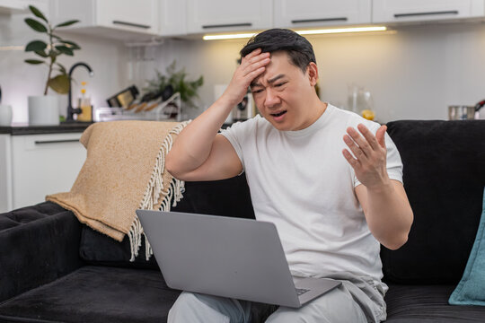 Asian man at home sees game loss on laptop touches forehead sighs and hits retry with focus. Chinese guy on sofa plans rematch sets target adjusts settings breathes and gets ready calm again steady
