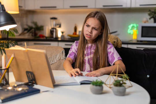 Teenager girl at night home reads problem from book for homework feels confused holds head sighs. Caucasian child kid sitting at table at evening puts notebook aside looks tries to understand slowly.