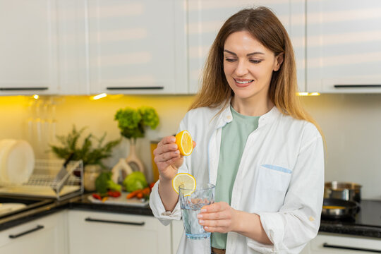 Middle-aged woman squeezing lemon into water in home kitchen for detox hydration and diet target. Young adult girl smiling while preparing healthy vitamin drink for weight loss and wellness routine.