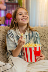 Young child girl eats popcorn and watches funny cartoon on television while sitting on sofa,...