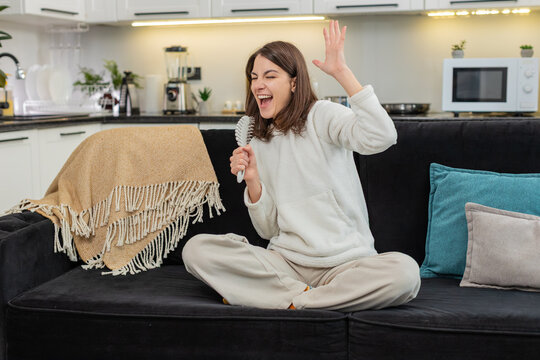 Young woman at home sings into comb like microphone dances and smiles with bold energy. Caucasian girl on sofa waves hand points at camera lip syncs chorus and enjoys music dream and star mood big joy