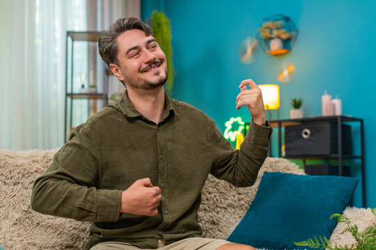 Middle-aged man at home rocks imaginary guitar strumming riff moving body enjoying big rock sound. Young adult guy on sofa performs wild air guitar solo active jumps celebrating music thrill passion