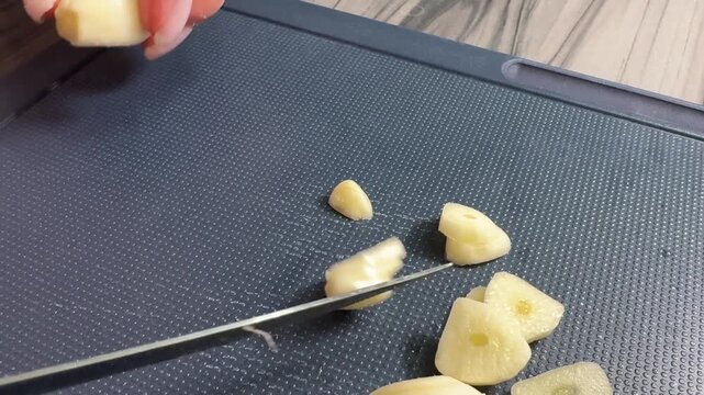 Close-up of a woman's hand slicing garlic cloves with a sharp knife on a wooden board, releasing pungent aroma from fresh white pieces.