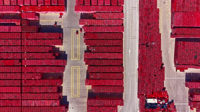 The scene shows a large number of red cargo containers arranged neatly in rows at a shipping port. Vehicles move along the paths separating sections of containers during the day.