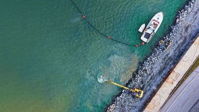 A small boat is anchored close to the shore while a machine works along the water's edge. The water is clear and the ground has rocks. This scene shows construction at a coastal site.