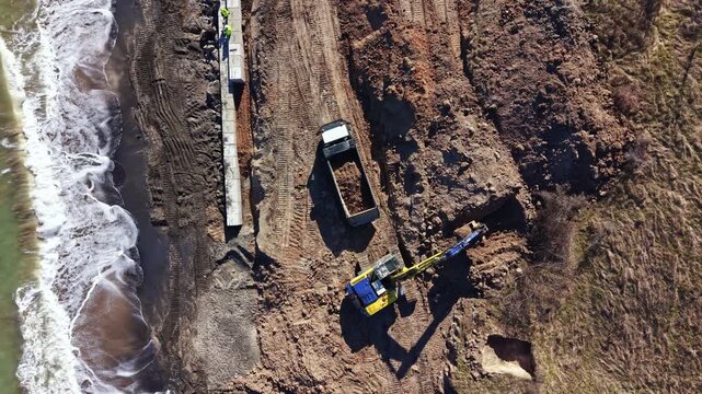 Heavy machinery is working on land excavation at a construction site located by the shore with visible waves and dirt being moved on a sunny day.