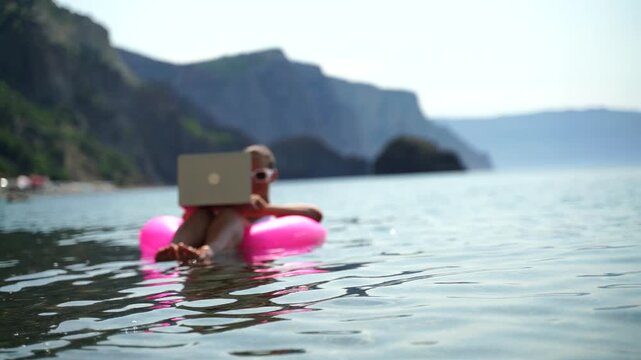 Woman works on laptop in sea. Freelancer, blond woman in sunglases floating on an inflatable big pink donut with a laptop in the sea at sunset. People summer vacation rest lifestyle concept.