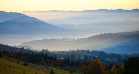 Naklejka premium Morning sunlight streams through mist, illuminating serene mountain village nestled among forested hills. Light creates ethereal rays that gently touch autumn foliage and houses scattered on slopes.