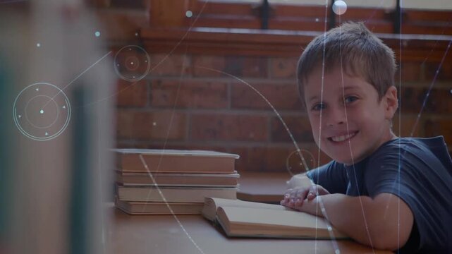 Male student sitting school desk reading open book looking up at camera prompt smiling amid bokeh