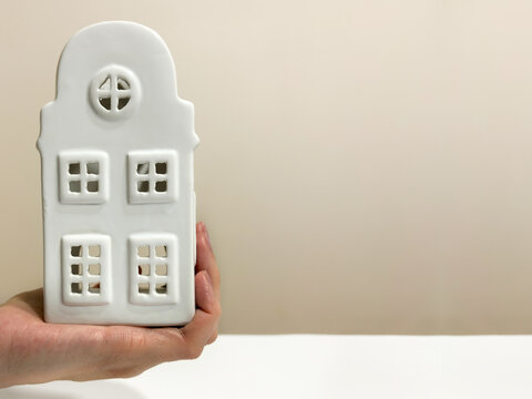 A hand holds a white ceramic house model over a table, representing real estate, home insurance, or mortgage concepts, with copy space