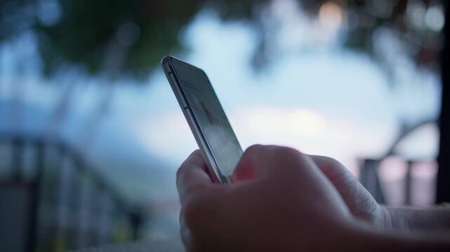 Close-up of male hands holding smartphone with stock market app on screen. Financial charts and trading interface visible while daylight and tree silhouettes create calm outdoor business atmosphere.