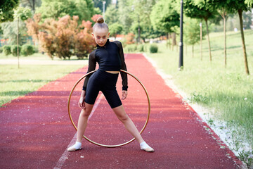 Fototapeta premium Young gymnast practicing rhythmic moves using a hula hoop on an outdoor track surrounded by greenery. Young Gymnast Practicing Rhythmic Routine Outdoors With Hula Hoop on Track