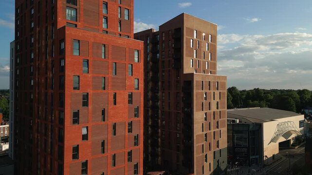 Modern brick apartment towers stand tall against a blue sky, with clouds and a hint of green trees in the background.