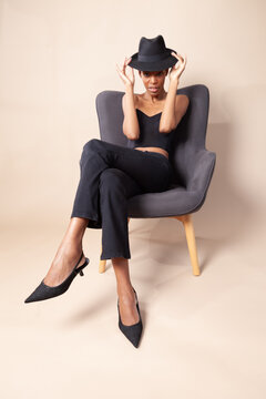 Playful black woman with short hair posing with fedora hat on armchair in photography studio