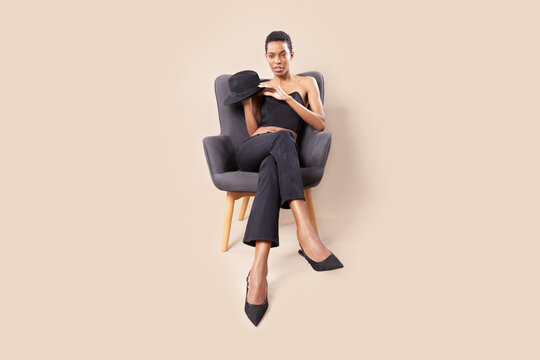 Playful black woman with short hair posing with fedora hat on armchair in photography studio