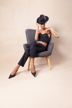 Playful black woman with short hair posing with fedora hat on armchair in photography studio