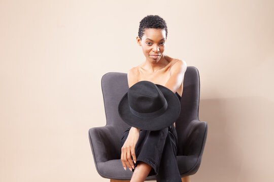 Playful black woman posing with fedora hat on armchair in photography studio
