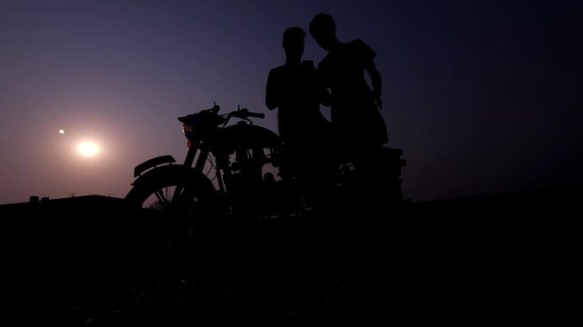 Silhouette of a motorcycle rider standing beside bike during sunset in India. 