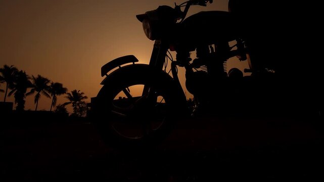 Silhouette of a motorcycle rider standing beside bike during sunset in India. 