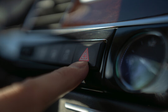 Finger presses hazard warning light button on car dashboard, activating emergency signal system in vehicle interior.