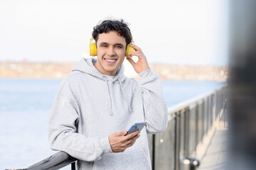 Fototapeta premium Young man in grey hoodie with headphones and mobile phone on embankment