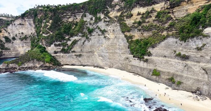 Drone view of Diamond Beach, Nusa Penida, Bali, featuring the iconic rock formation, white sand shoreline, turquoise waves, and limestone cliffs with lush greenery. A world-famous tropical destination