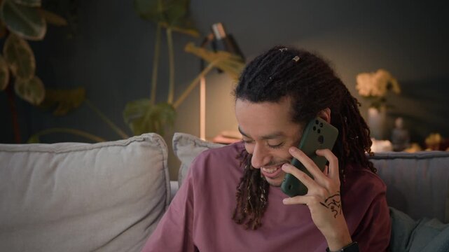 Tilt medium shot of positive young multiethnic man calling on mobile phone while working on wireless laptop and taking notes at home