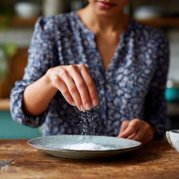 Female asian adult sprinkling salt into plate at wooden table in kitchen casual blouse