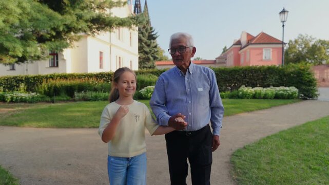 Grandfather reminisces during walk. Older man recounts family tales while strolling. Seasoned grandfather exchanges heritage stories with his granddaughter during peaceful park walk