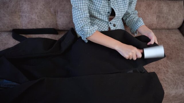 Meticulous woman sitting on a brown sofa using a sticky lint roller to remove dust, hair, and lint from a black coat, ensuring her clothes are perfectly clean before wearing