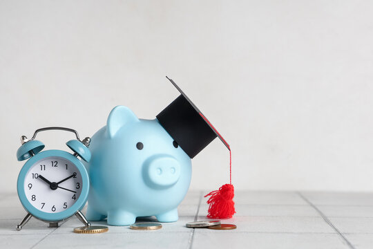 Piggy bank with graduation hat, money and alarm clock on white tile table near wall. Tuition fees concept