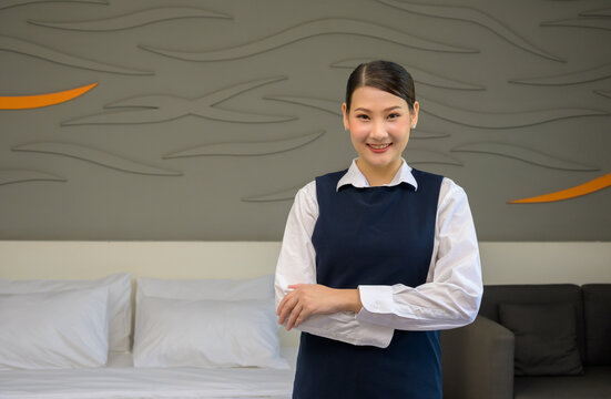 Confident smiling young female hotel housekeeper in uniform standing with arms crossed in modern luxury bedroom or resort guest suite