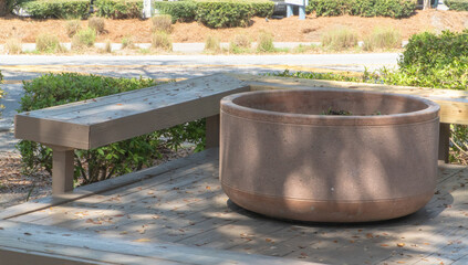 Naklejka premium Large round concrete planter next to a weathered wooden bench on a deck with fallen leaves, illuminated by natural sunlight in a peaceful outdoor setting, perfect for relaxation.
