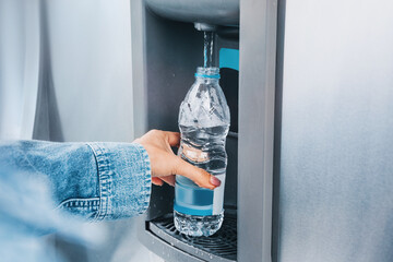 Person's hand holding a recyclable plastic bottle, filling it with clean drinking water from a modern dispenser for hydration © EdNurg