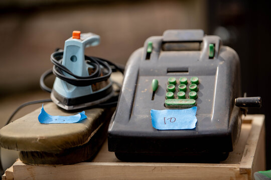 An old cash register and a cord-wrapped iron tagged with blue painter's tape prices sit ready for sale, a documentary image of estate sales and vintage secondhand culture and outdated technology. 