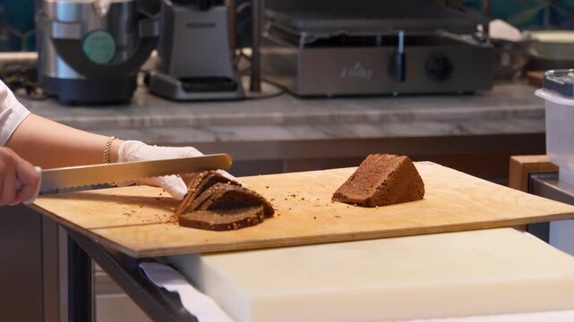 Professional Baker Slicing Dark Rye Bread with Seeds. Chef slicing a triangular loaf of dark coriander rye bread on a wooden board. Professional kitchen setting for bakery, restaurant