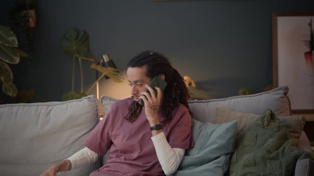 Medium long shot of young multiethnic businessman with dreadlocks calling on smartphone, working on wireelss laptop and taking notes while sitting on couch in living room, copy space